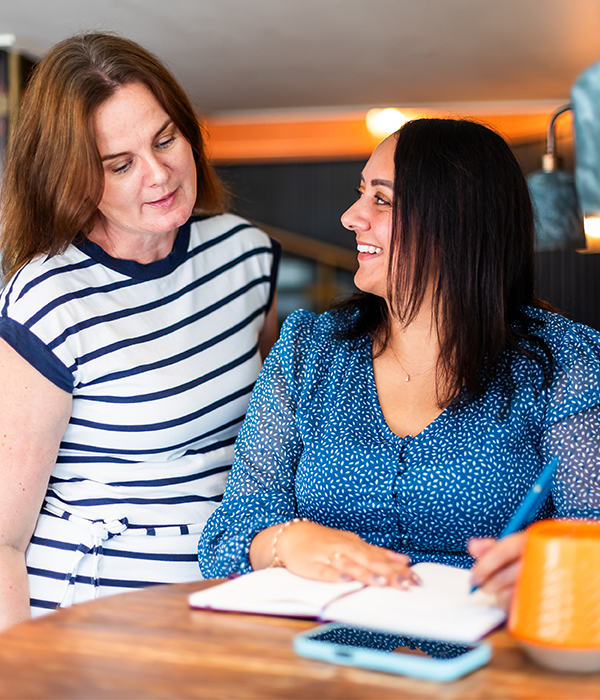 Emma Culver looking over Bianca Rodriguez Jacobs' shoulder at writing pad on desk.