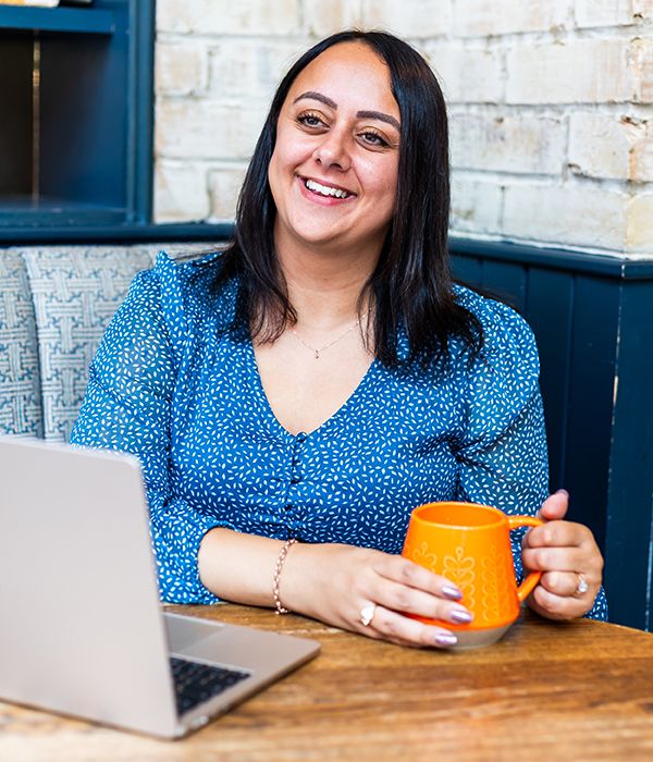 Bianca Rodriguez Jacobs smiling with orange mug sitting at table with open laptop.
