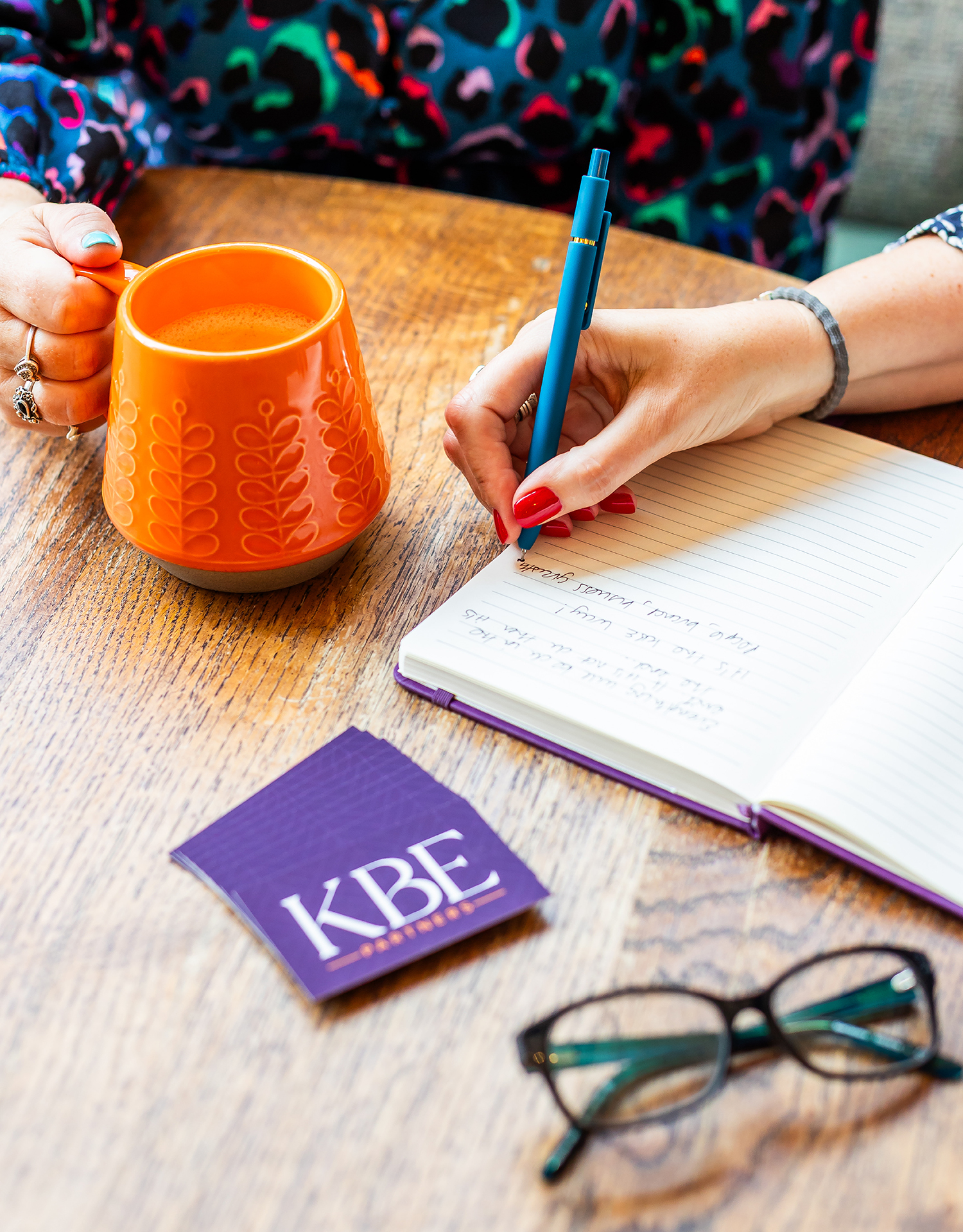 Overhead view of desk with female hand with red finger nails writing in journal with orange mug, pair of glasses and KBE Partners Business cards on desk.